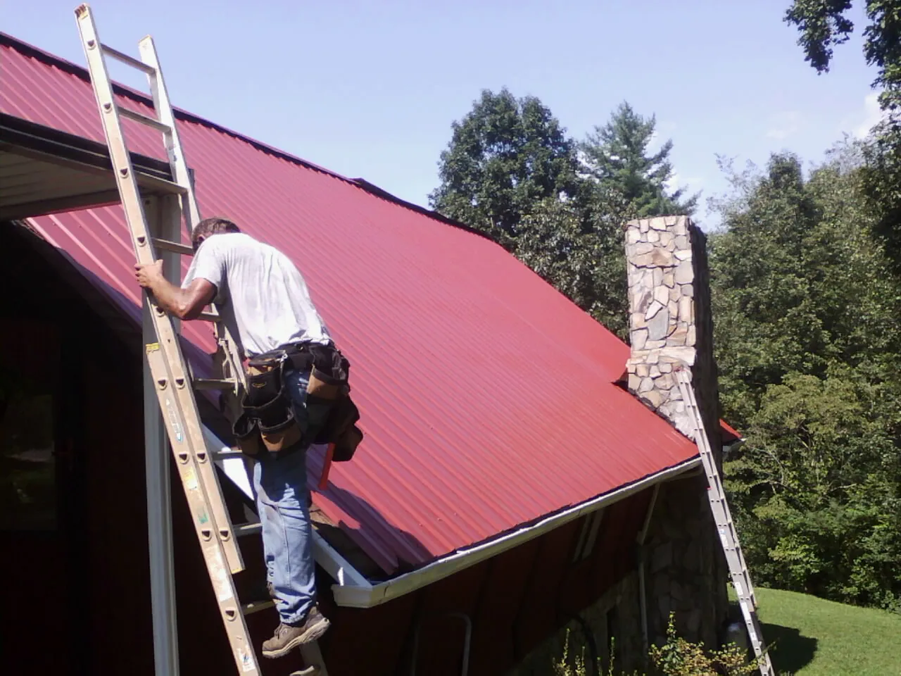 Roofer on a ladder beside a red metal roof and stone chimney — Soffit & Fascia Repair by Roofing Brothers in Sterling Heights